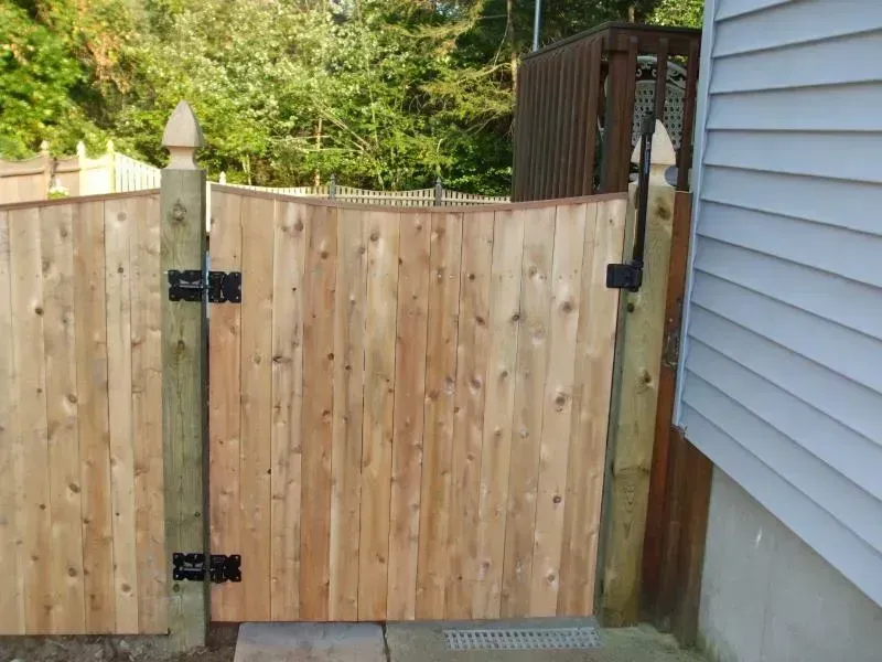 A wooden gate with black hinges and a latch, attached to fence posts next to the side of a light-colored house.