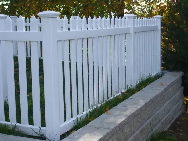 A white vinyl picket fence installed atop a gray stone retaining wall outdoors with green trees in the background.