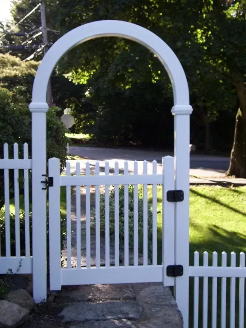 A white picket fence featuring a matching gate topped with a large, rounded archway in a sunny outdoor setting.