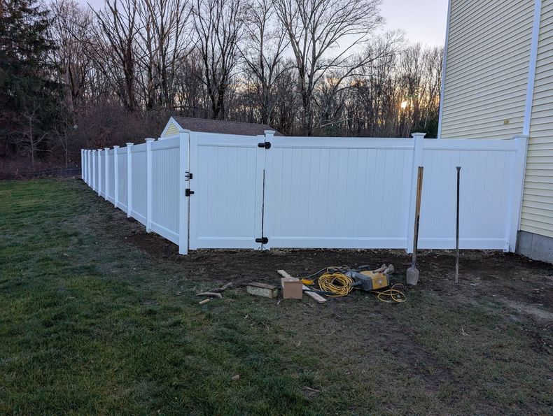 A newly installed white vinyl privacy fence with a gate, situated on a grassy lawn next to the side of a yellow house.
