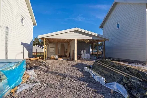Construction site with unfinished carport between two white houses; gravel ground, blue sky.