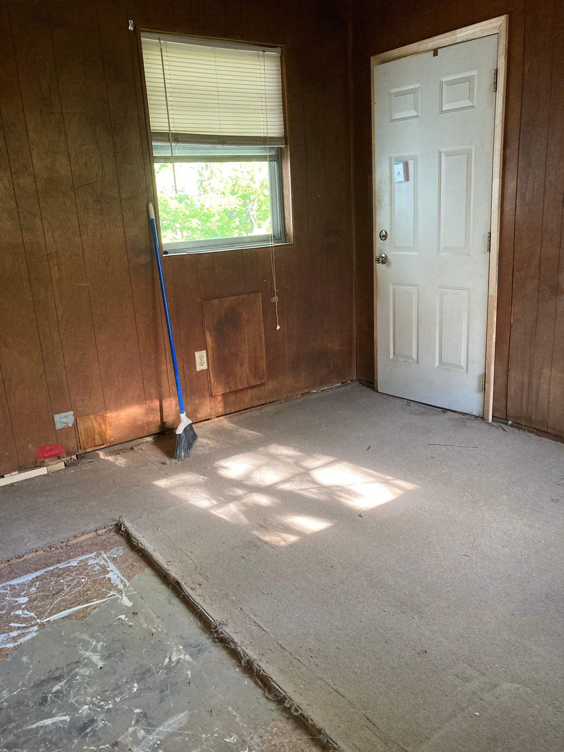 Interior room with brown paneling, a white door, a window with blinds, and a broom.