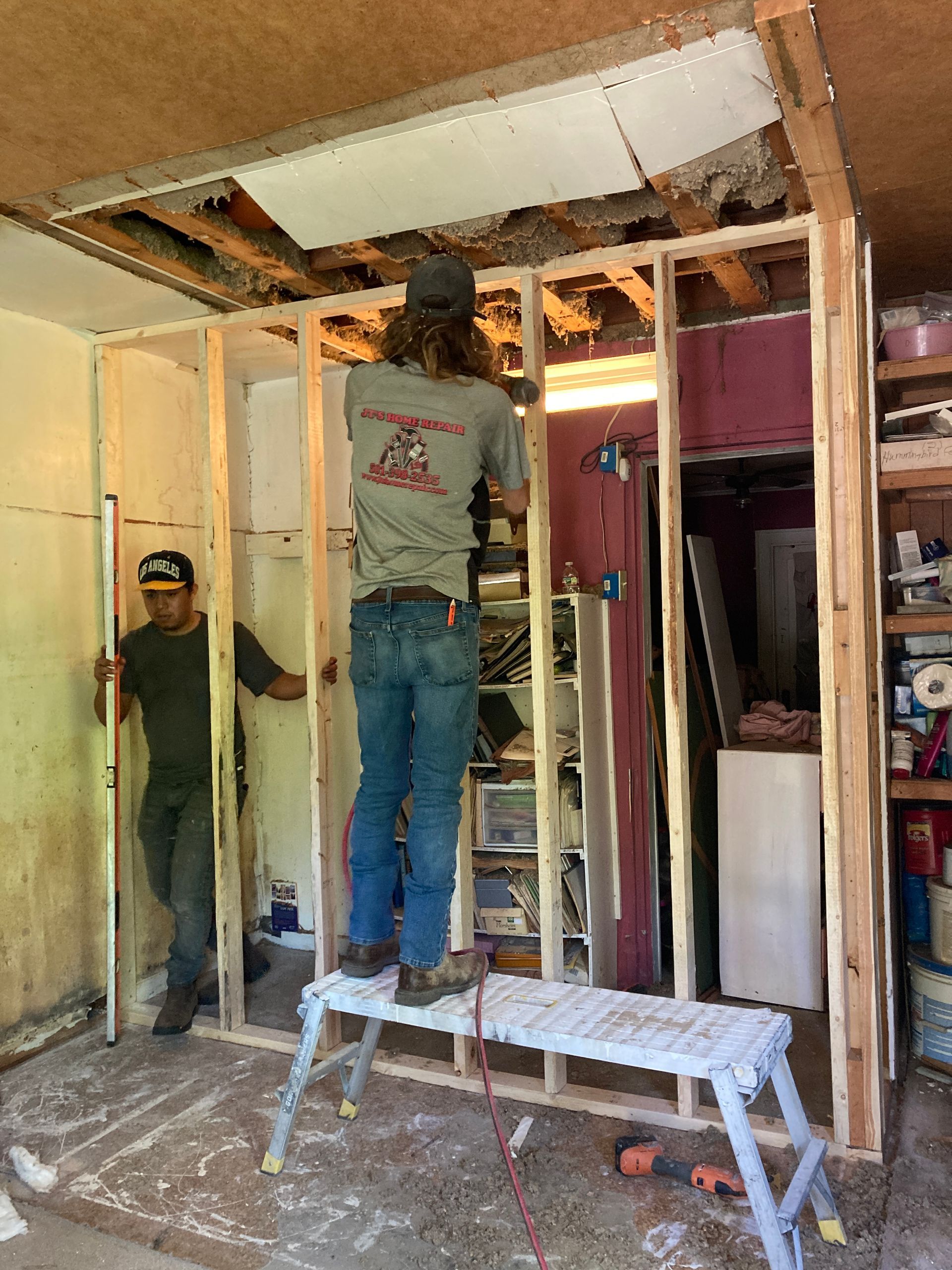 Two men building a wall inside a room. One on a stepstool, the other holding a level.