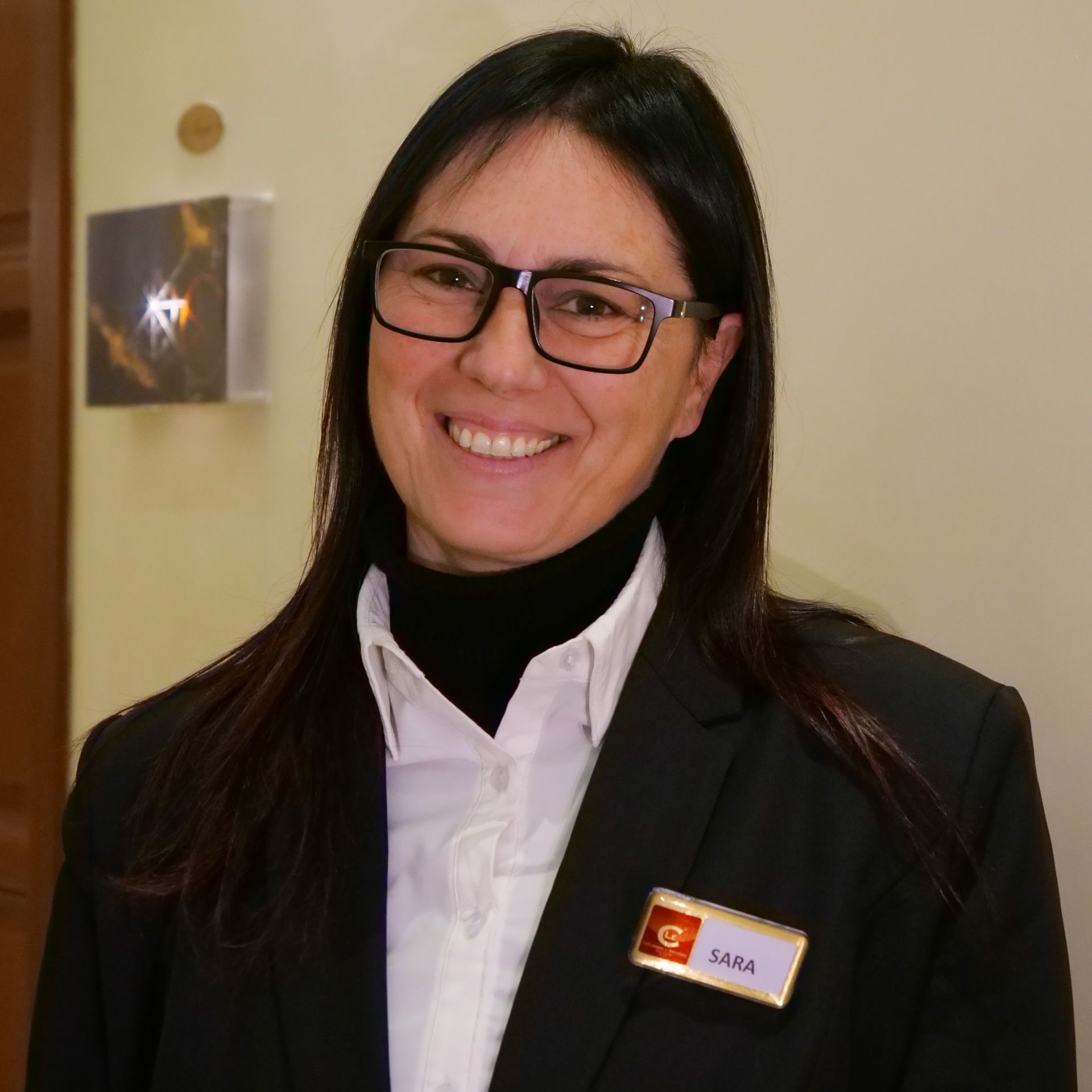 Woman in a black blazer and glasses smiles, wearing a name tag.