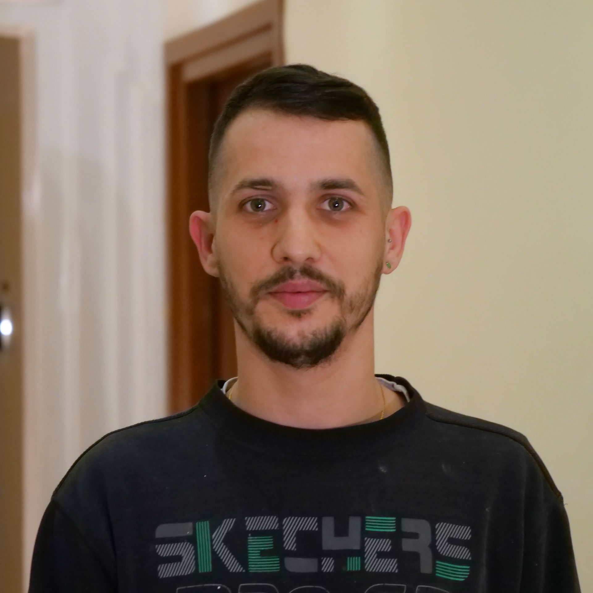 Man with short dark hair and a beard, wearing a black sweatshirt, standing indoors.