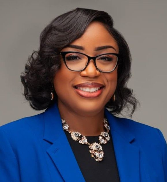 Woman with glasses, wearing blue blazer and statement necklace, smiling.