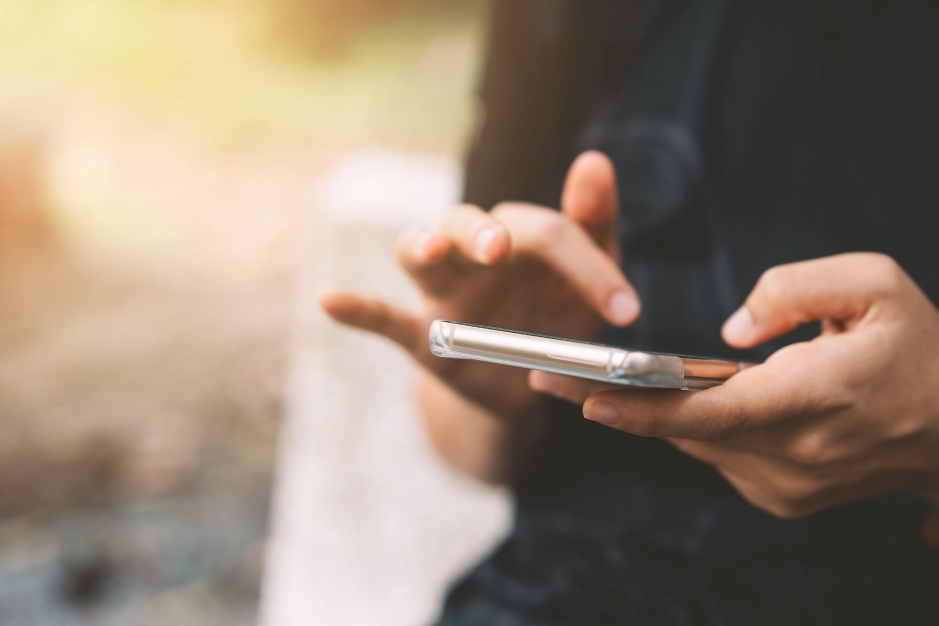 A person's hands holding a smartphone, with one finger touching the screen in a soft, sunlit setting.