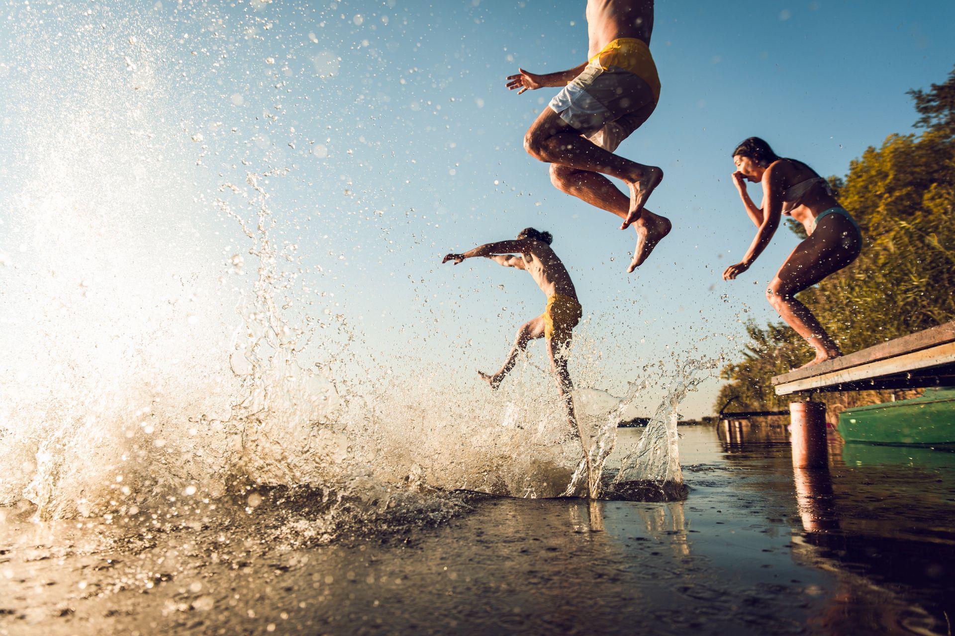 Three people jumping into water from a wooden dock, creating a large splash; sunny setting.