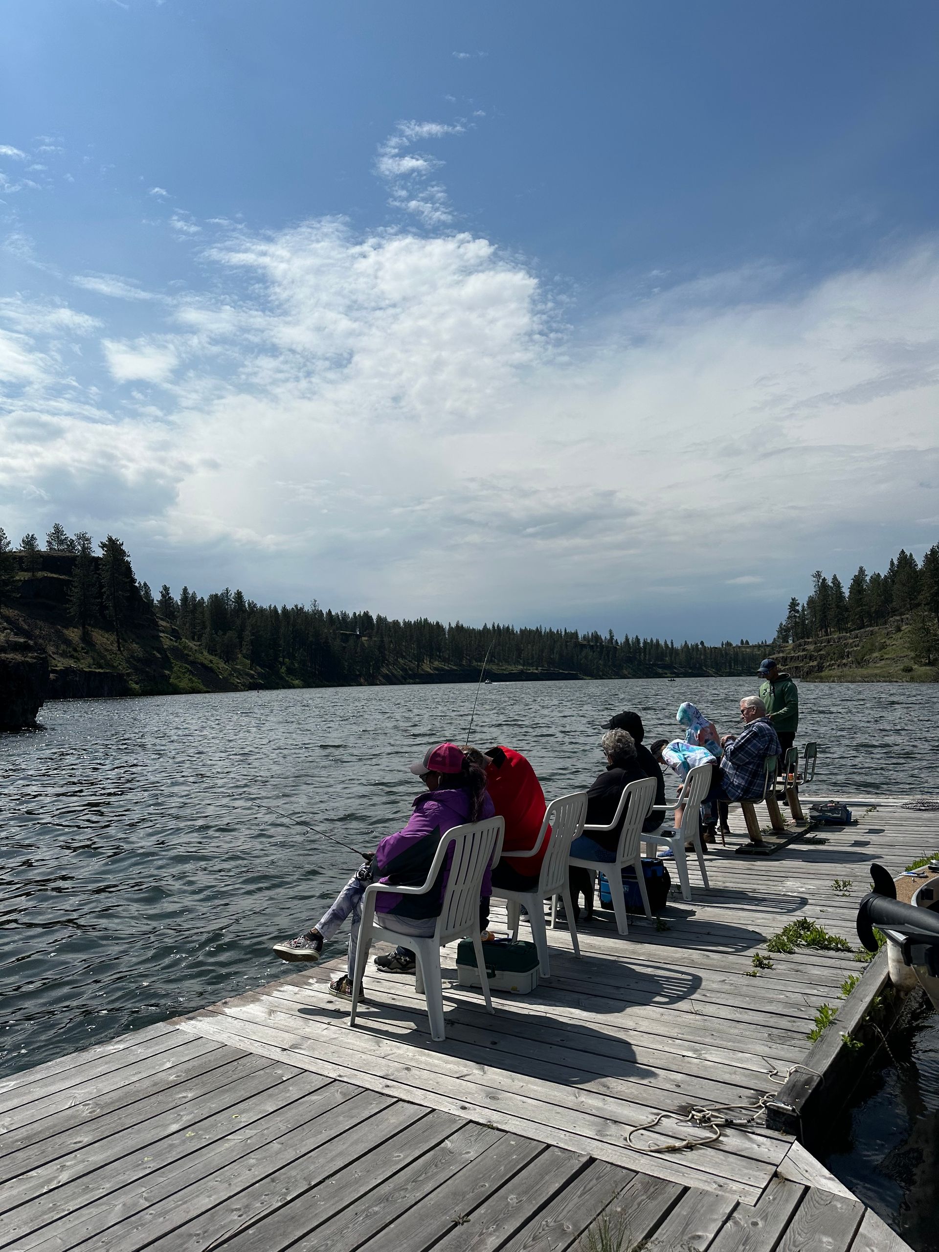 People sitting on chairs on a wooden dock, overlooking a lake with trees under a partly cloudy sky.