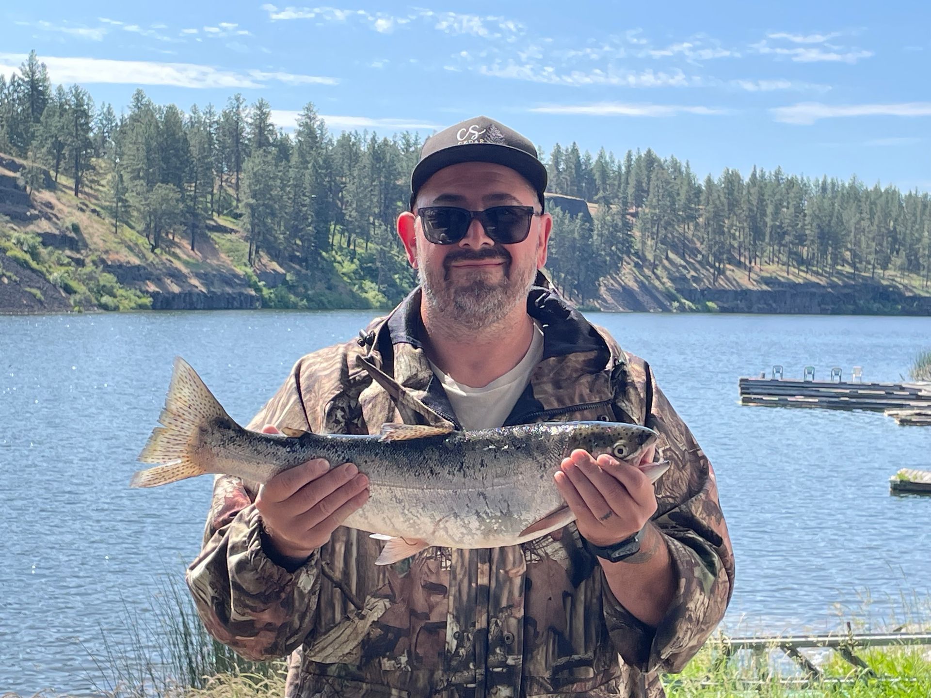 Man in camo holding a large fish by a lake, with trees in the background.