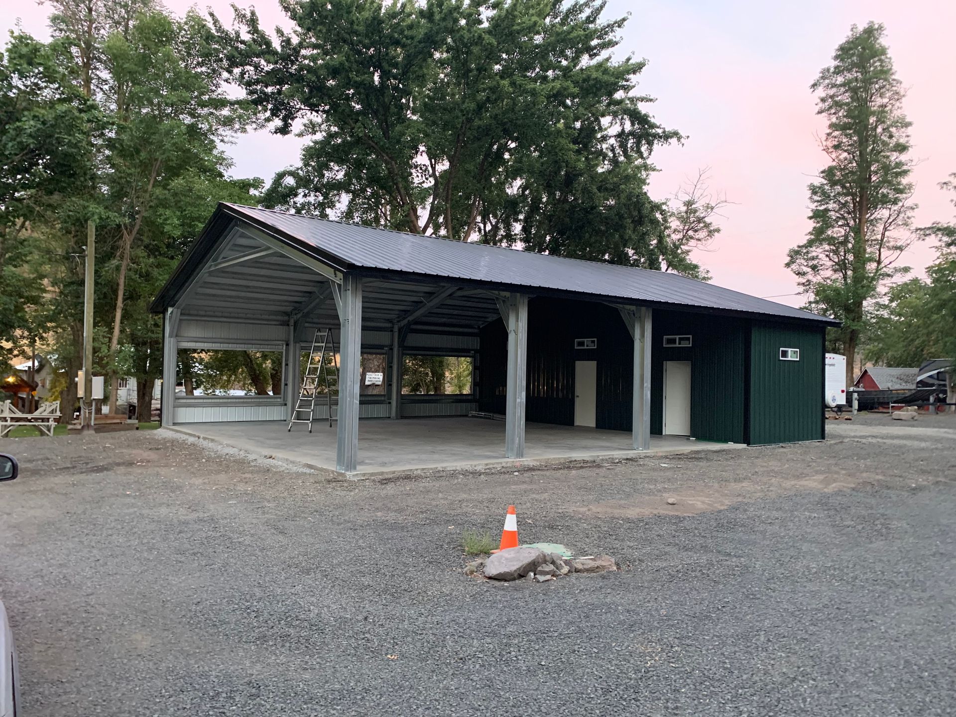 Open-air metal building with a green-sided enclosed section; gravel ground, trees, and an orange cone are visible.