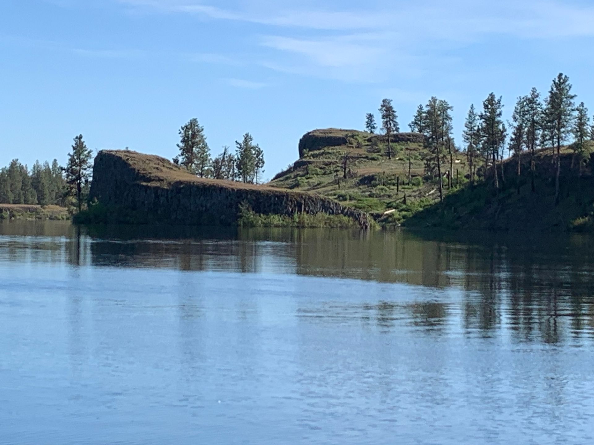 River scene with dark cliffs and sparse trees reflected in the water under a clear blue sky.