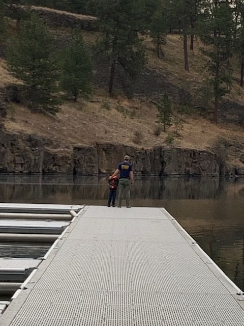 Man and child stand on a dock, looking toward the water, with a rocky hillside and trees in the background.