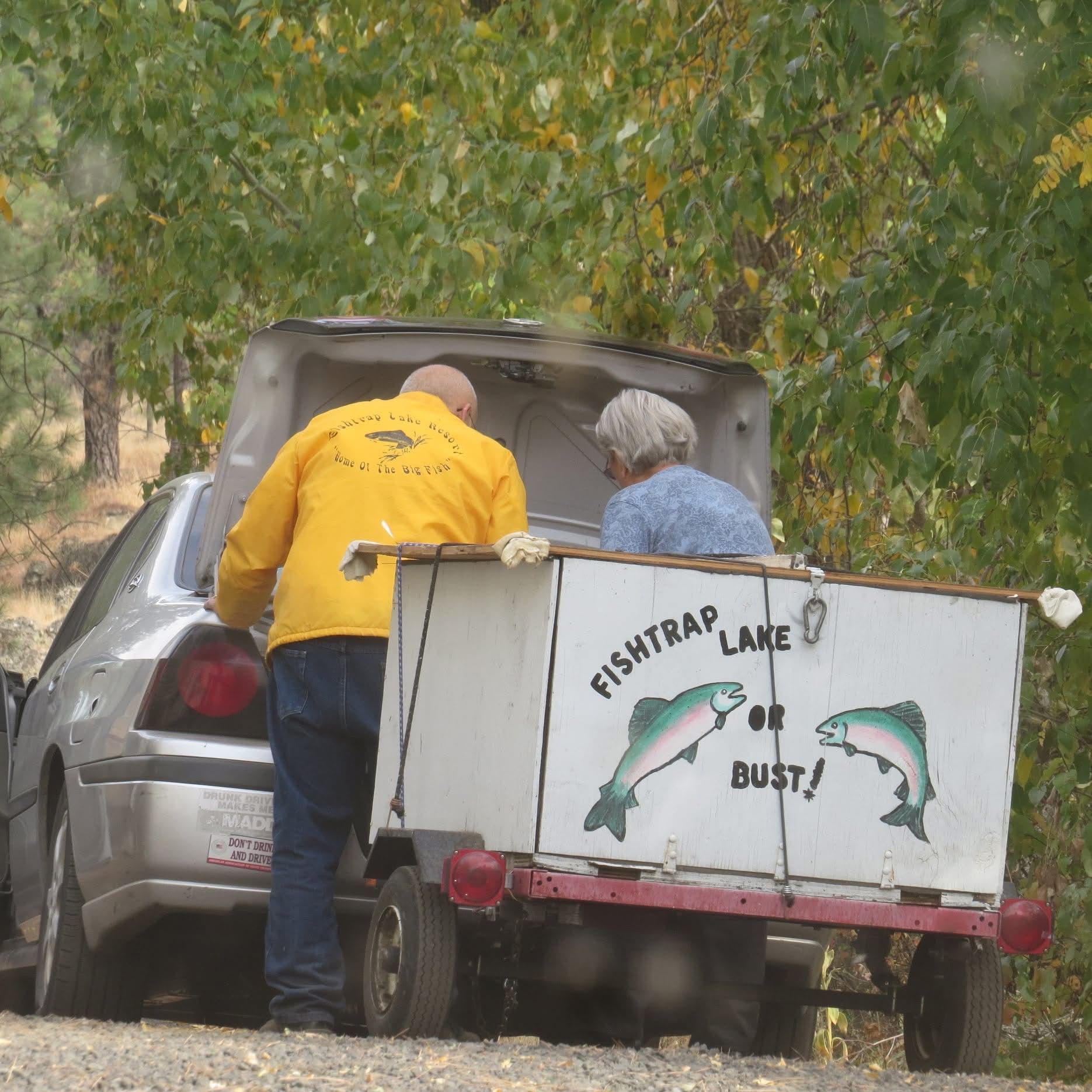 Man in yellow jacket and woman by car, hooking up a trailer that says 