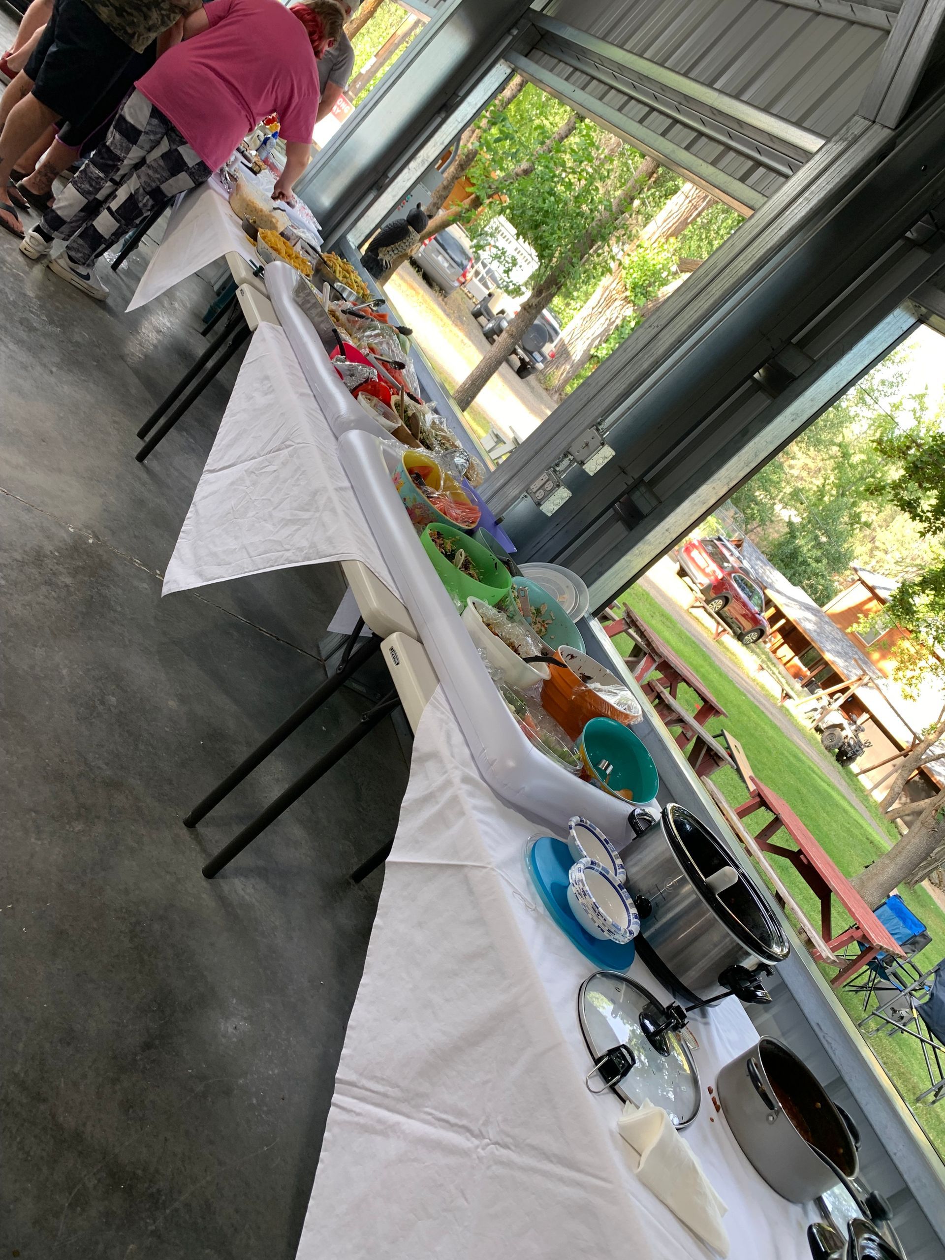 Long table with food dishes and pots, people standing near in an outdoor covered area.