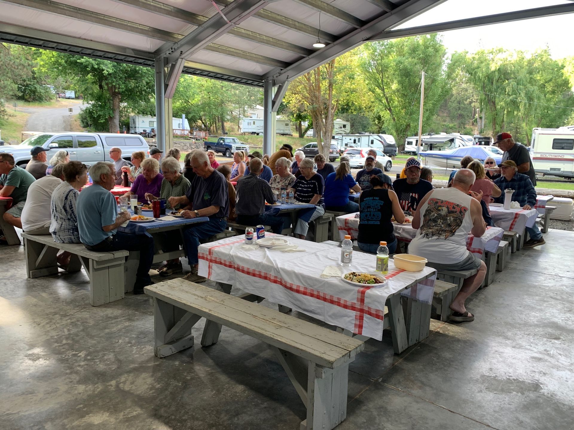 People seated at picnic tables under a covered pavilion, eating and socializing outdoors.