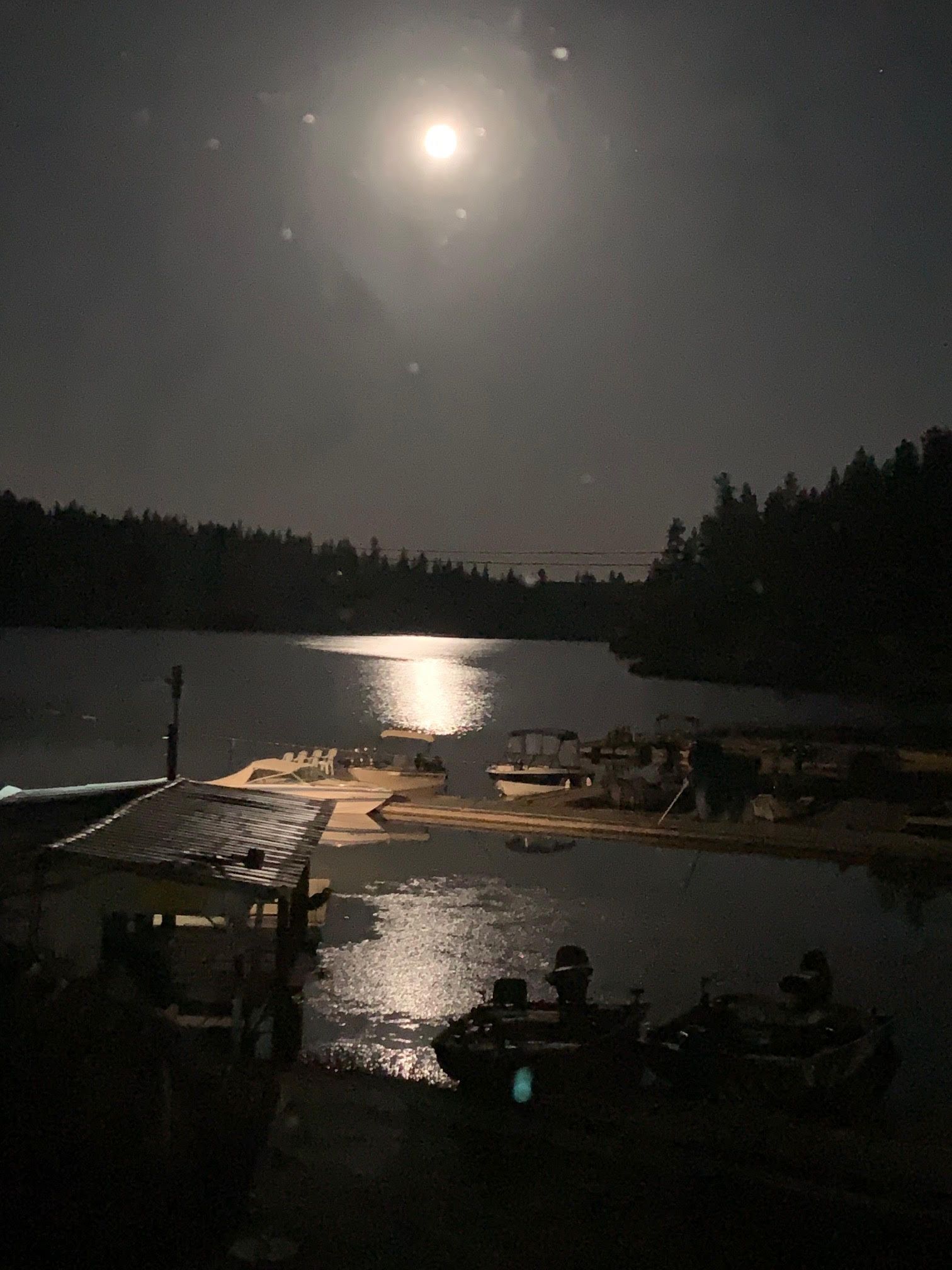 Full moon reflecting on a dark lake with a dock, boats, and treeline in the distance.