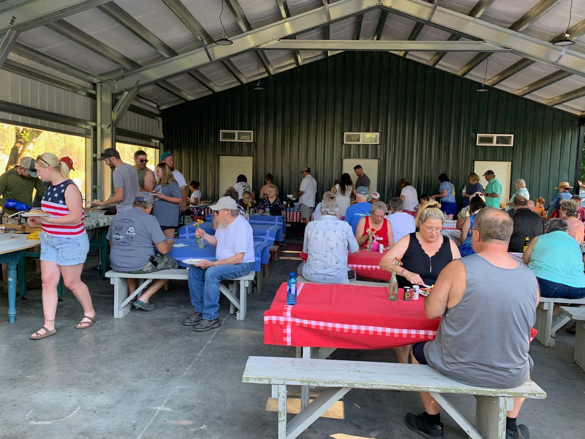People at a covered picnic area; some seated at benches, eating. Red, white, and blue tablecloths.