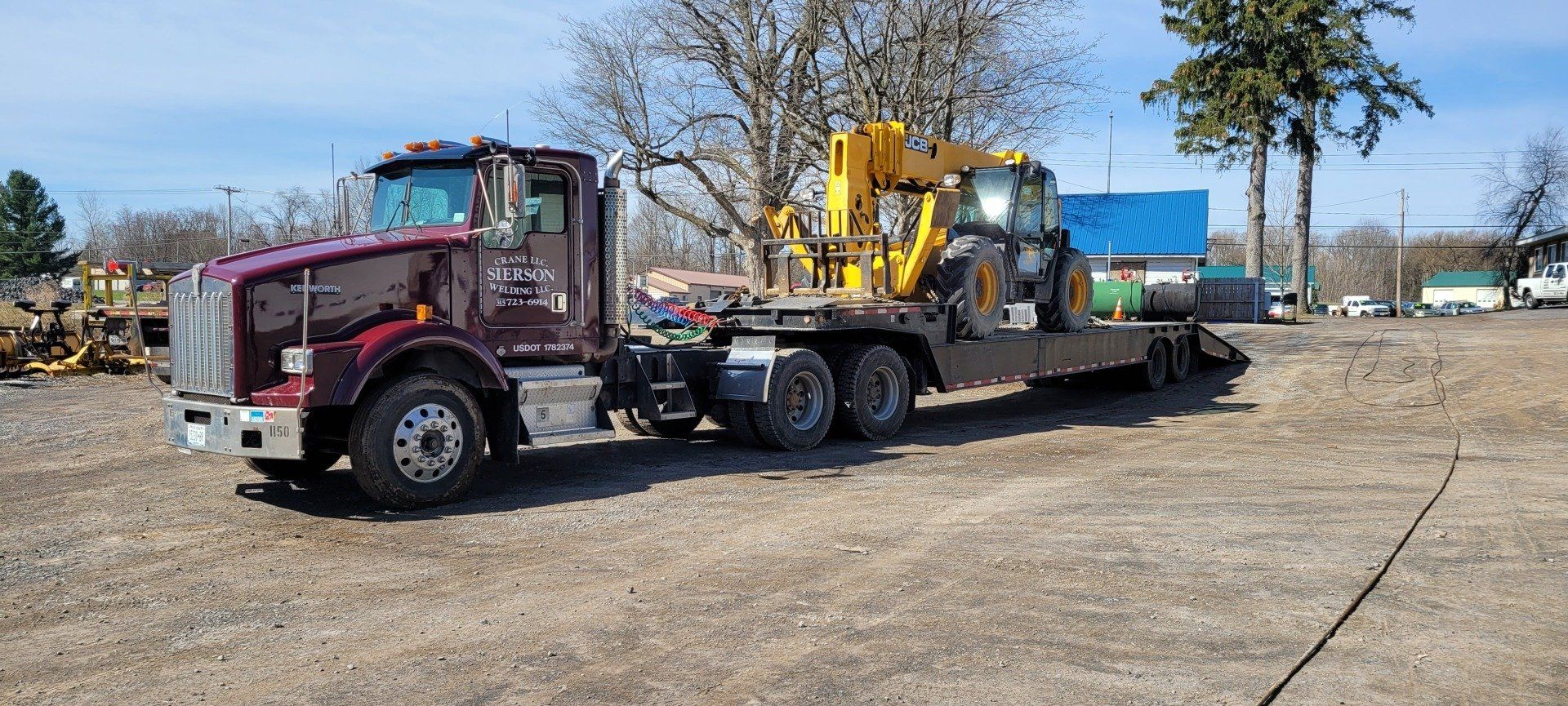 A maroon semi-truck hauling a yellow backhoe on a flatbed trailer, parked on gravel in a sunny area.