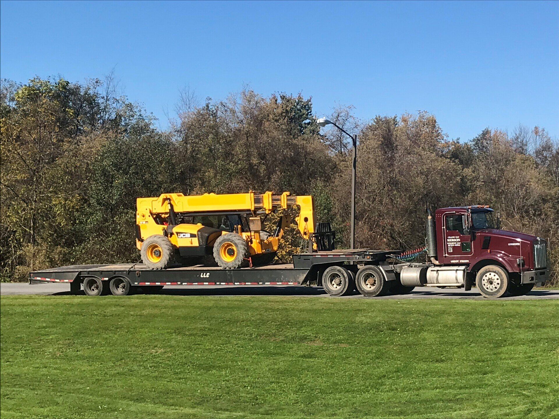 A yellow construction vehicle is being transported on a flatbed trailer by a maroon semi-truck on a grassy area.