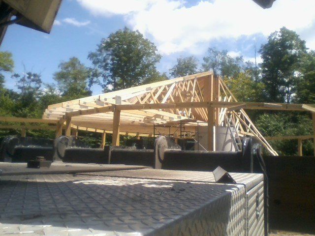 Construction of a wooden building's roof with visible trusses, set against a backdrop of trees and a bright sky.