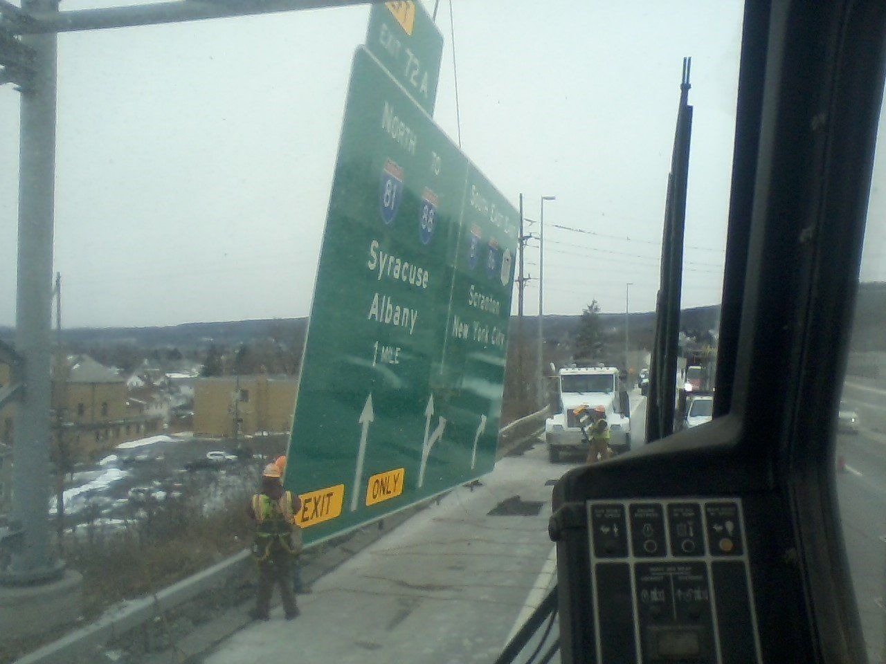 A large green highway sign being installed over a road by a crane.  Road crew in orange vest.