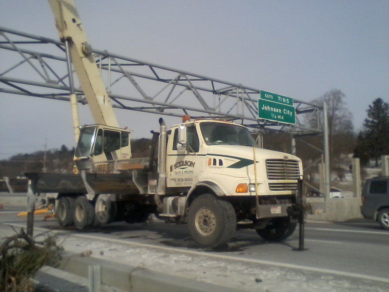 A large, cream-colored truck with a crane parked on a highway, under a road sign.