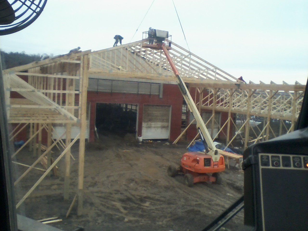 Construction site with workers on a roof, using a lift. Red building with wood framing, overcast sky.