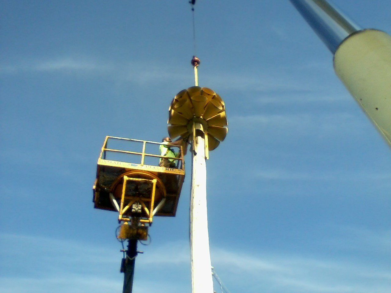 Crane lifts a large, yellow, circular structure toward a pole. A person on a lift platform assists. Blue sky backdrop.