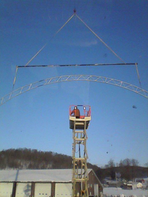 Crane lifts a metal arch over a person in a lift. Blue sky, snow, building in background.