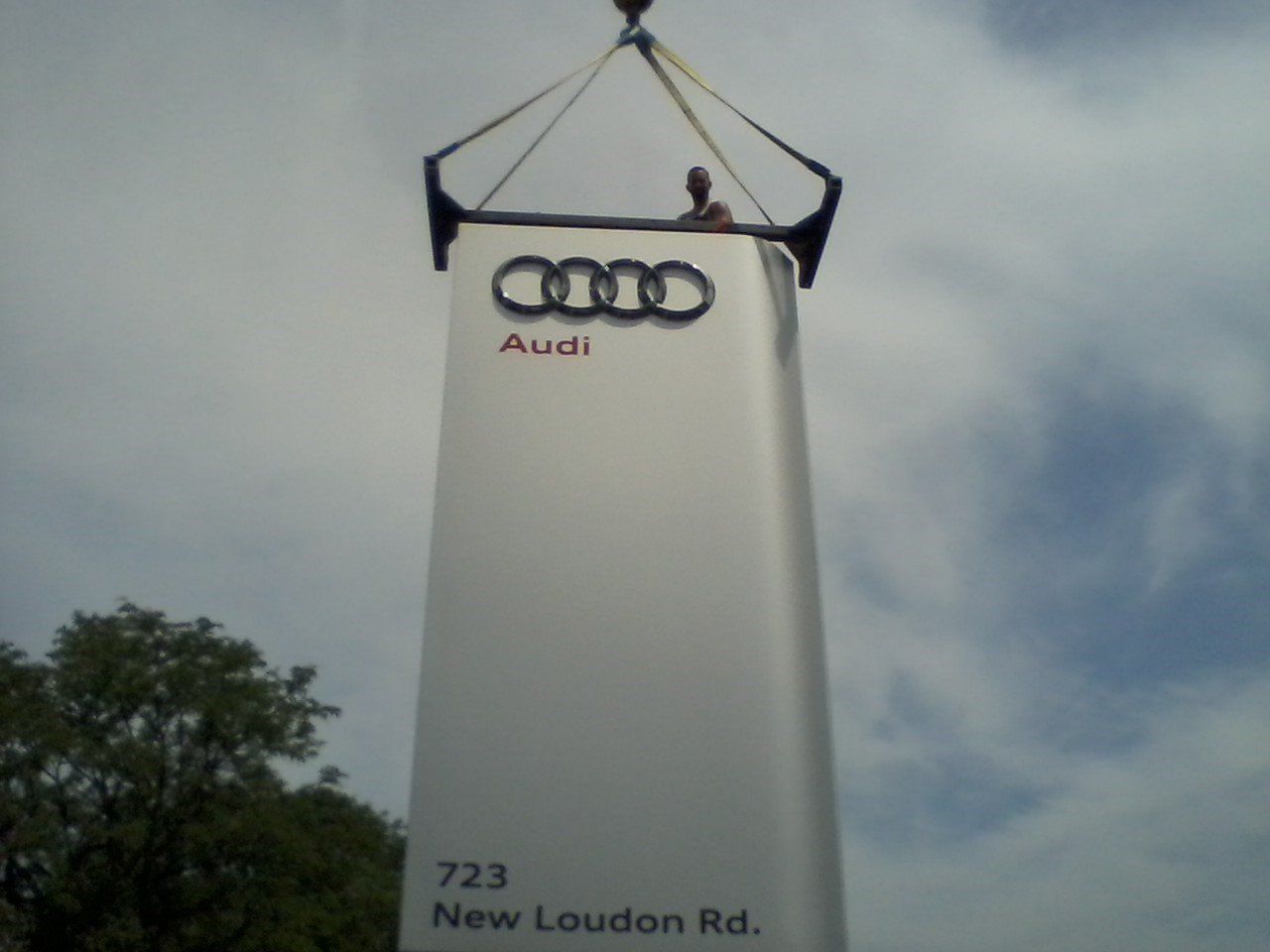 An Audi dealership sign being lifted into place by a crane. Includes the Audi logo, address, and person.
