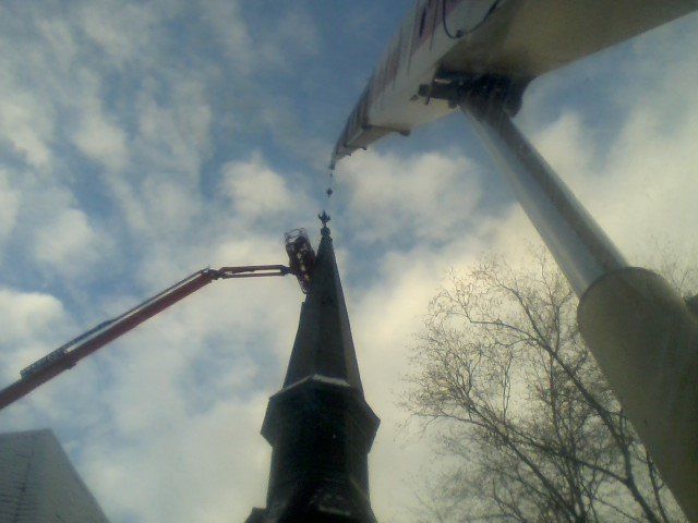 Workers on a lift repairing the steeple of a church against a cloudy sky.