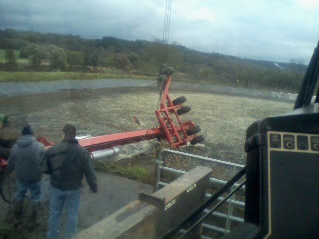 Two men look at a tipped-over red farm implement near water in a field on an overcast day.