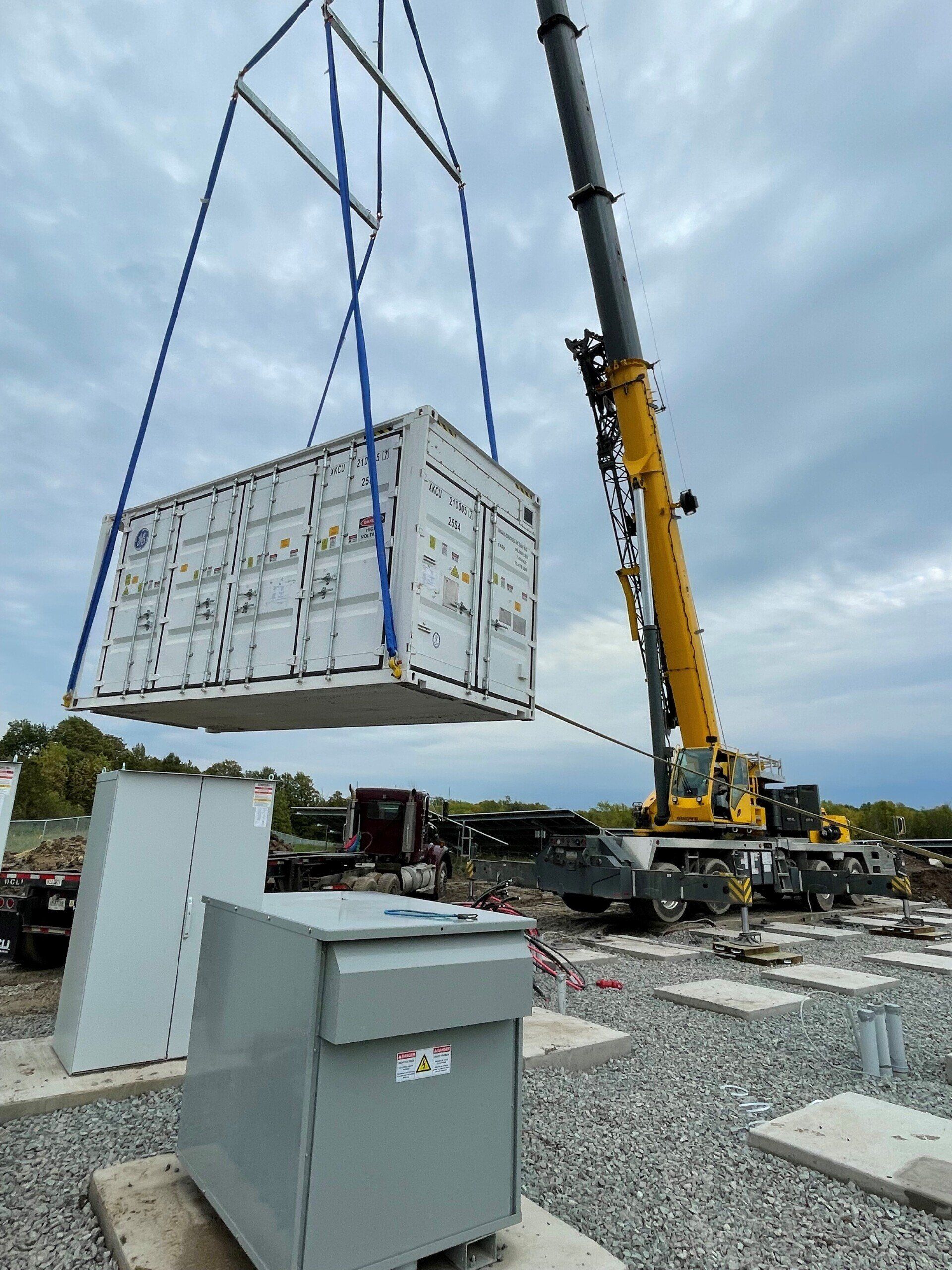 Crane lifting a white container over a gray electrical box at a construction site.
