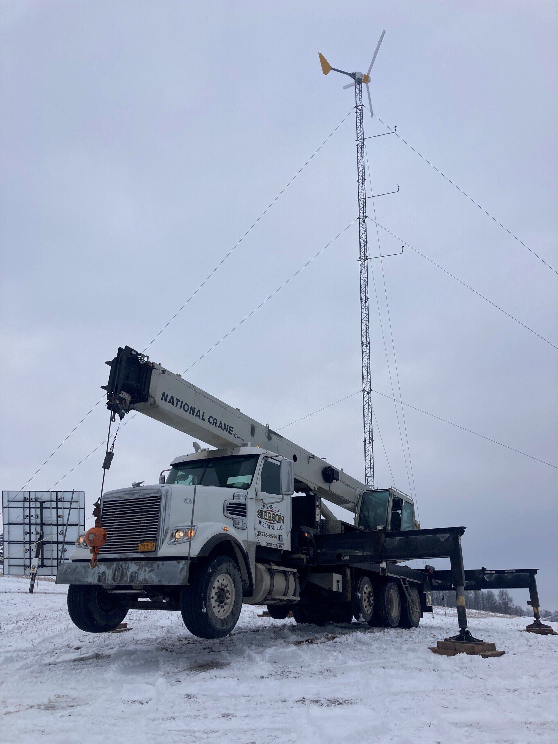 A crane truck installing a small wind turbine in a snowy field on a cloudy day.