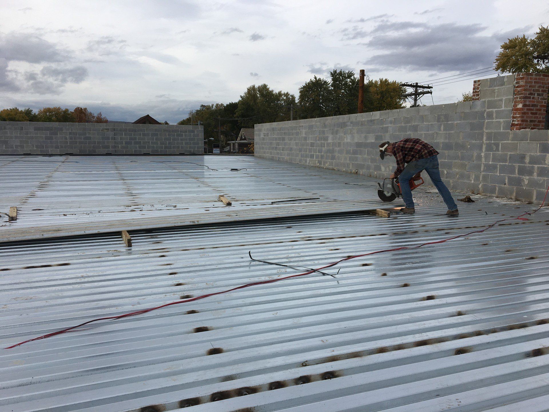 Construction worker hammering metal decking on a building rooftop. Gray metal, concrete blocks, cloudy sky.