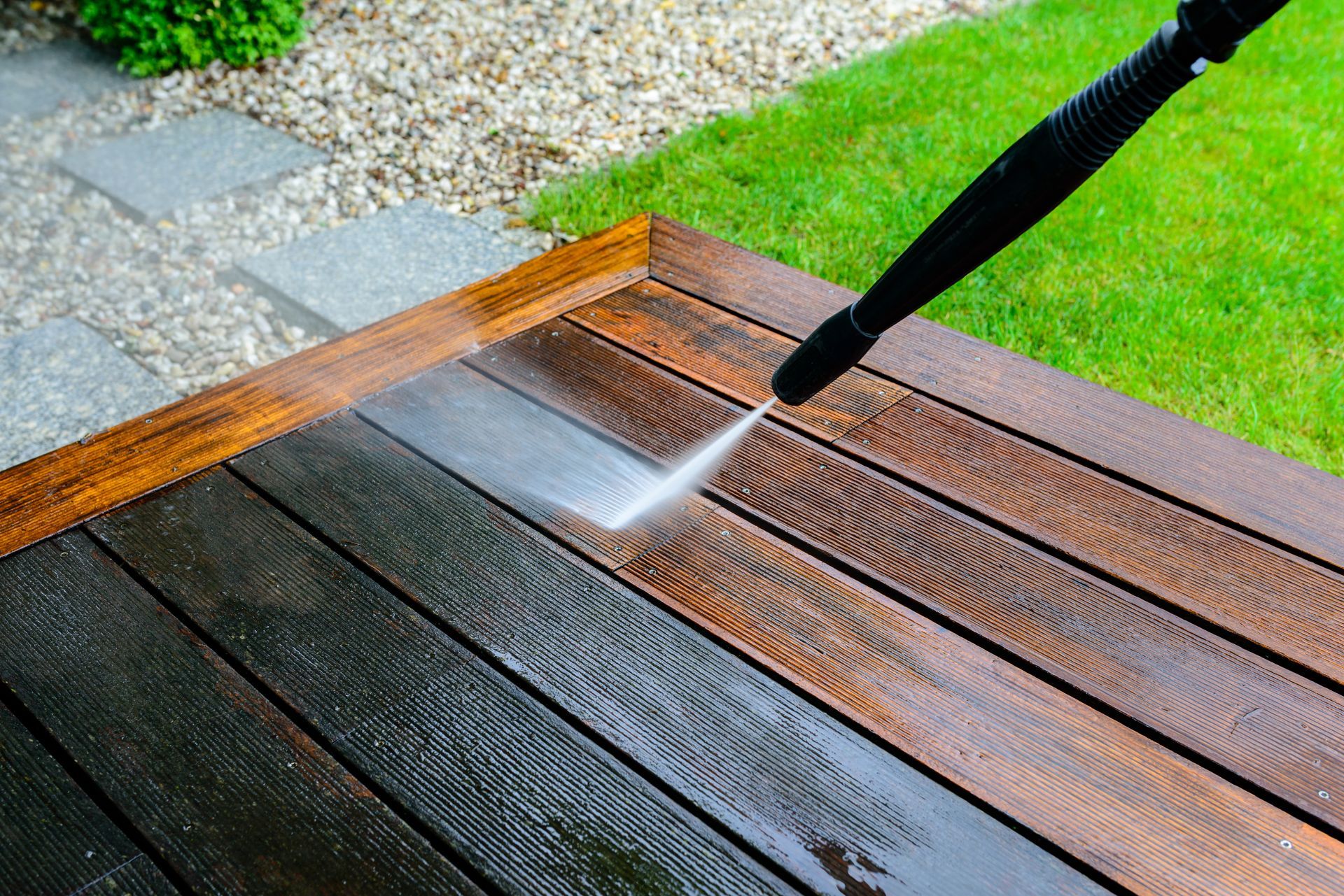 Pressure washer cleaning a wooden deck beside a gravel path and green lawn