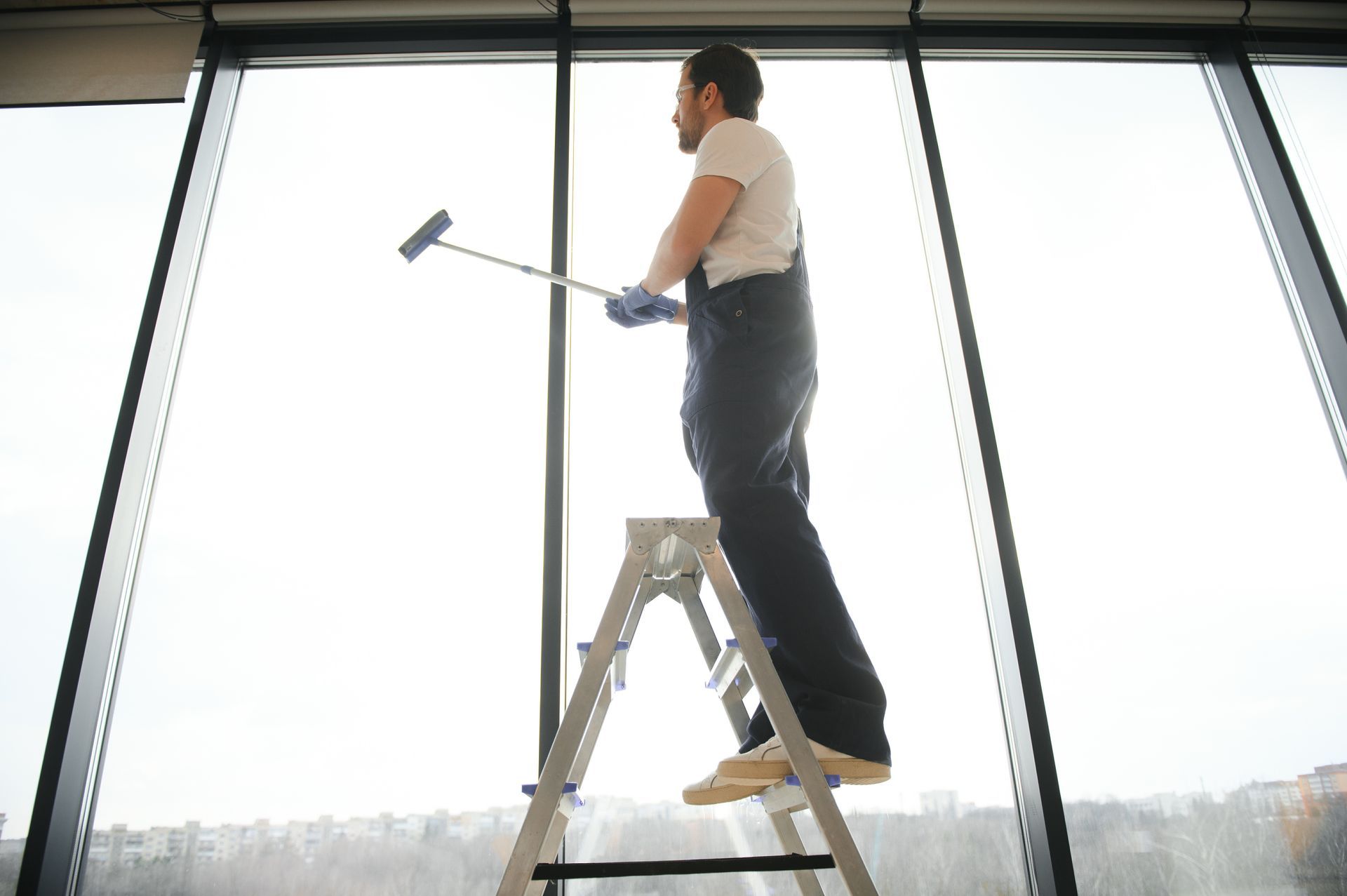 Person on a step ladder cleaning large windows with a squeegee in a bright room