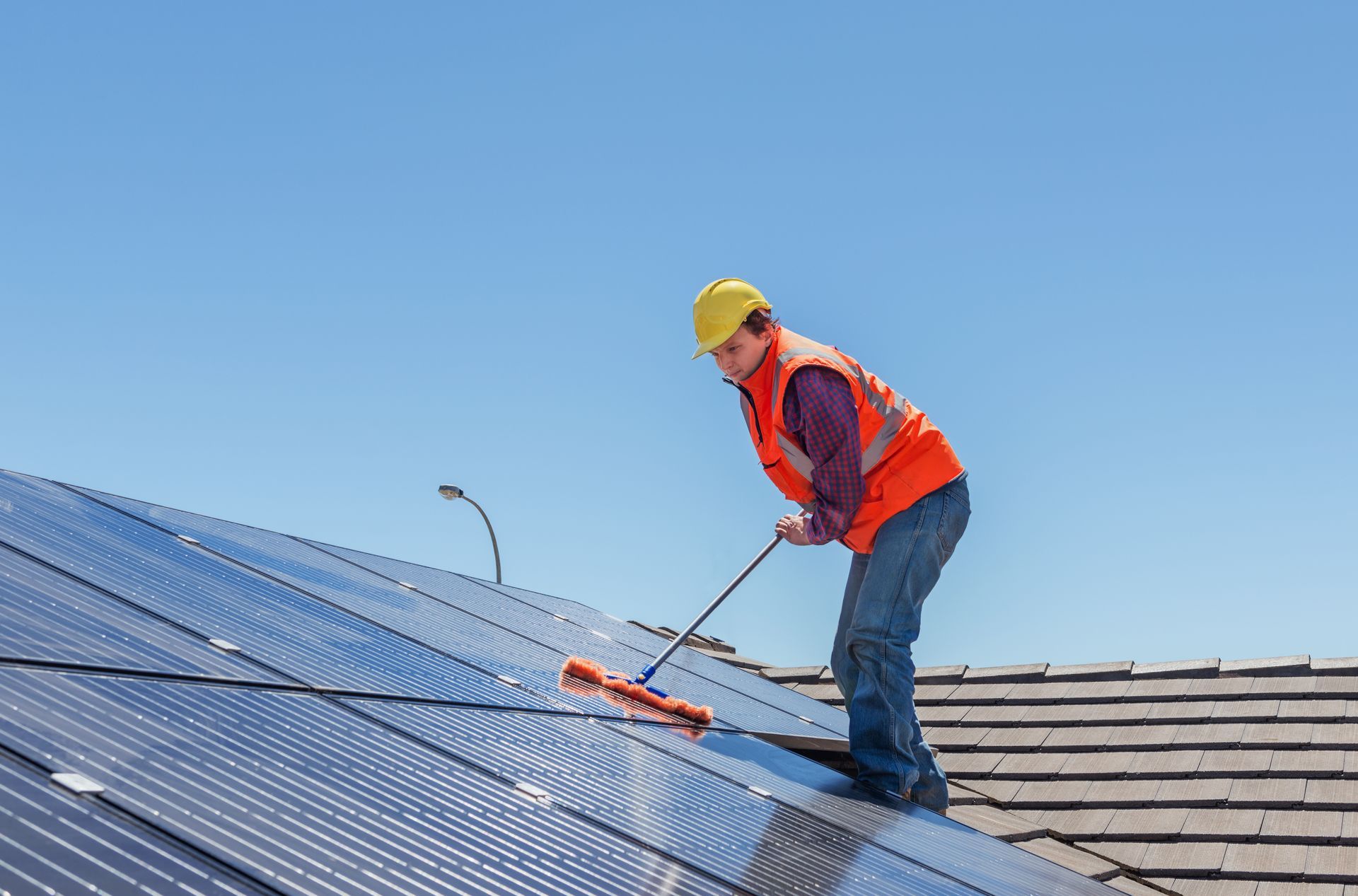 Worker in orange safety vest and yellow hard hat cleaning a rooftop with a long brush. Worker in orange safety vest and yellow hard hat cleaning a rooftop with a long brush.