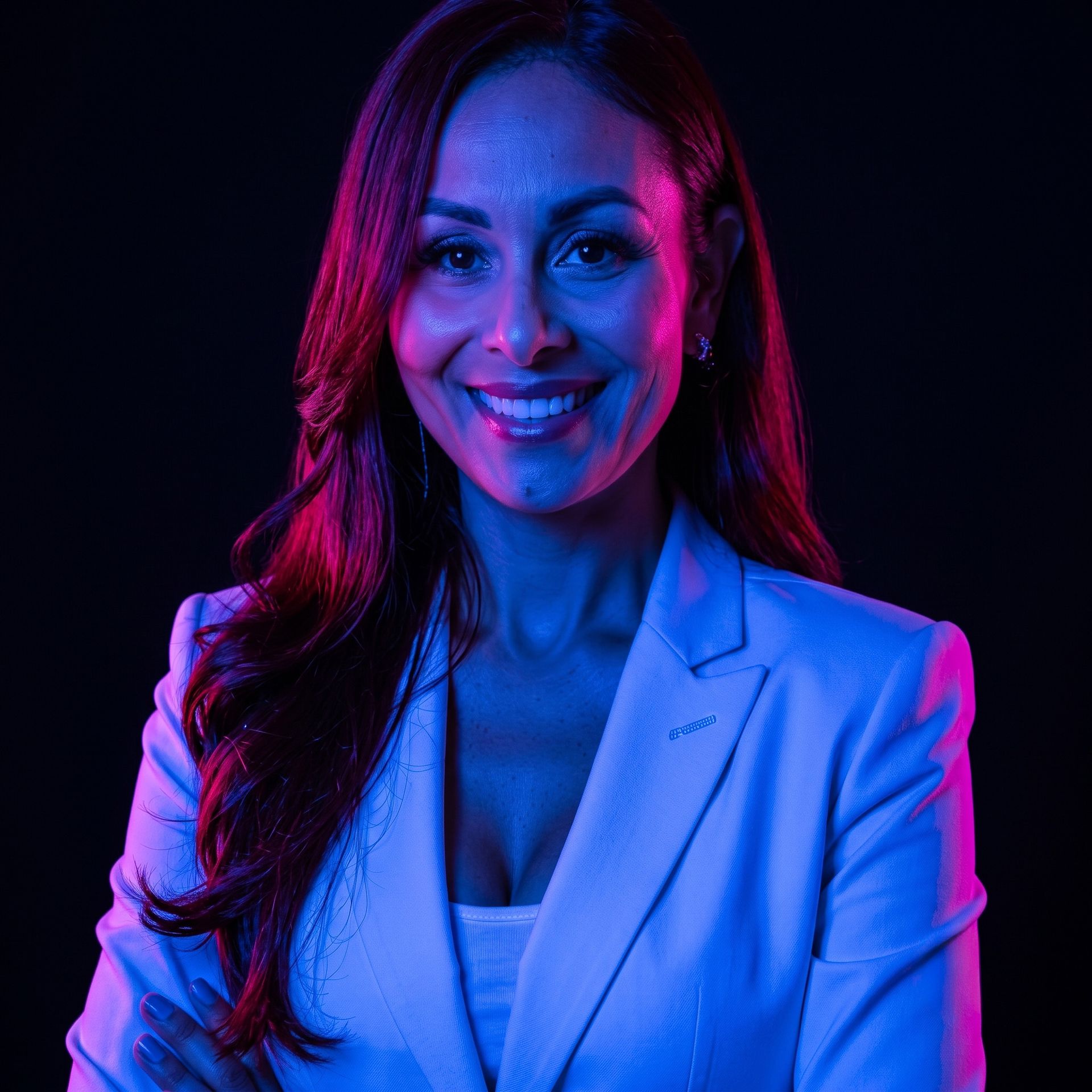 Portrait of a smiling woman in a light blazer, lit by vivid blue and pink neon against a dark background