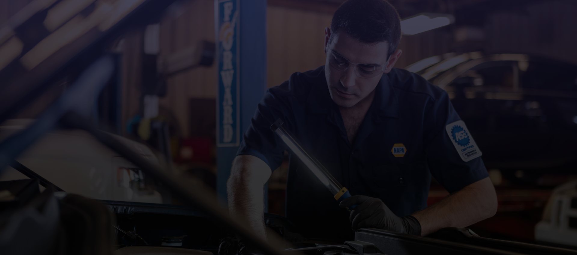 Mechanic working on a car in a garage, using a light to examine the engine. | Broadway Automotive