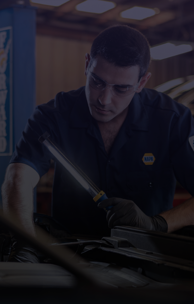 Mechanic in dark uniform uses a light to inspect a vehicle engine, shop setting. | Broadway Automotive