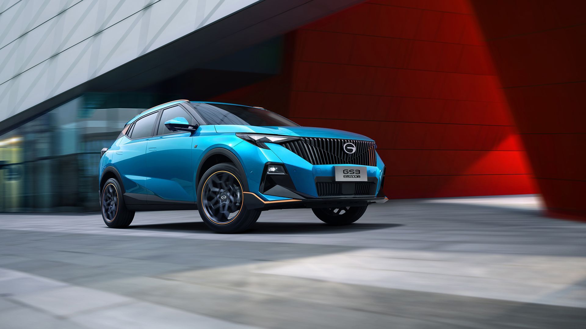 Um SUV azul brilhante passando em frente a um prédio moderno com uma parede vermelha e uma praça pavimentada.