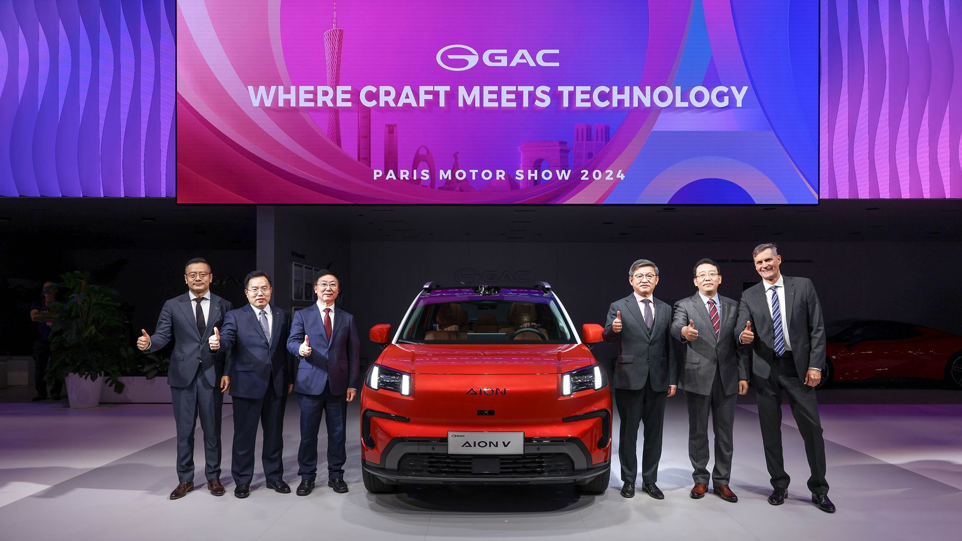 Seis pessoas de terno posam ao lado de um veículo GAC vermelho brilhante em um palco do Salão do Automóvel de Paris.