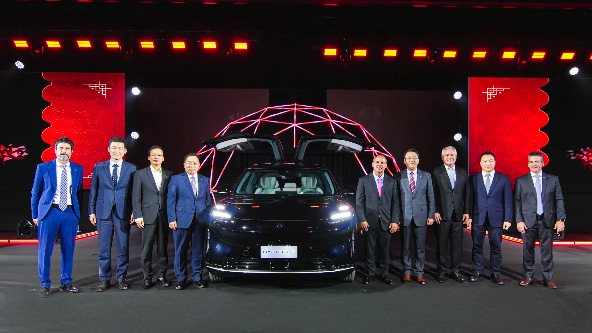 Um grupo de pessoas de terno em pé em um palco atrás de um veículo preto com as portas abertas em um evento em local fechado.