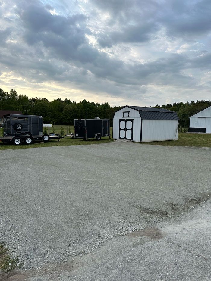A row of trailers are parked in a gravel lot next to a barn.