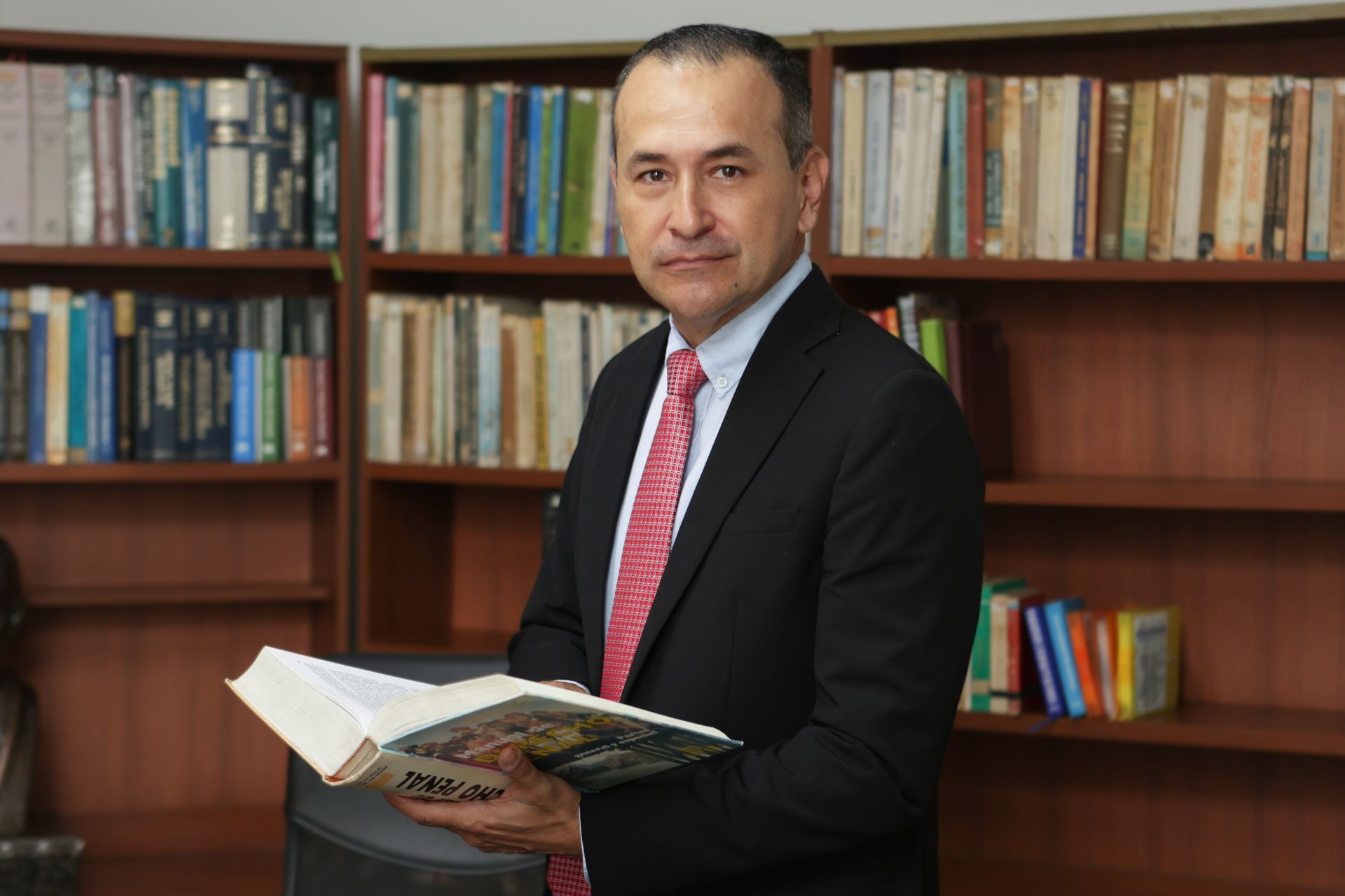 Hombre con traje sosteniendo un libro, de pie en una biblioteca.