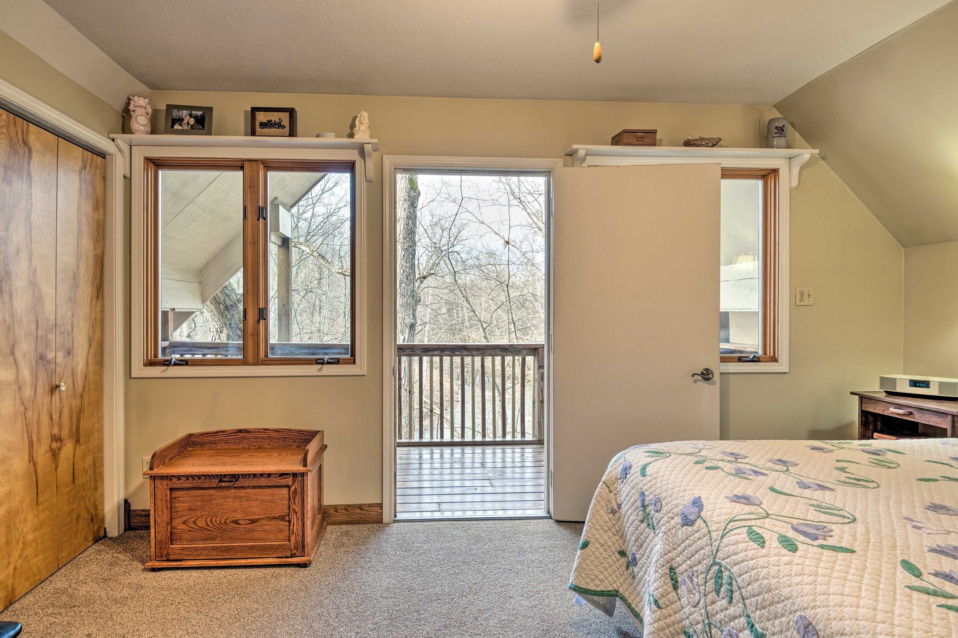 Bedroom with balcony door, window, and bed. Light brown carpet, wooden chest, and neutral walls.