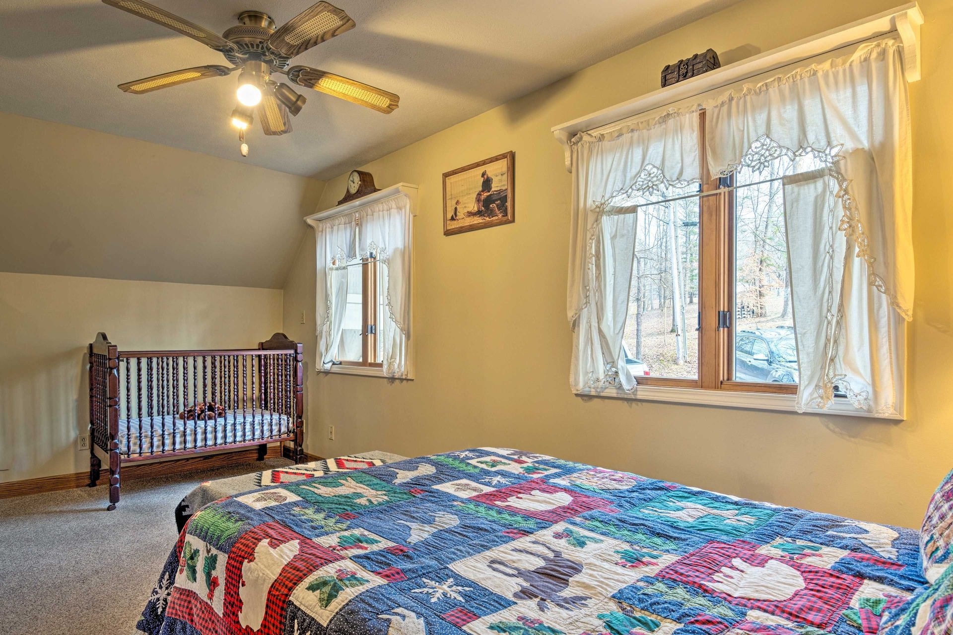 Bedroom with crib, bed with quilt, two windows with curtains, ceiling fan.