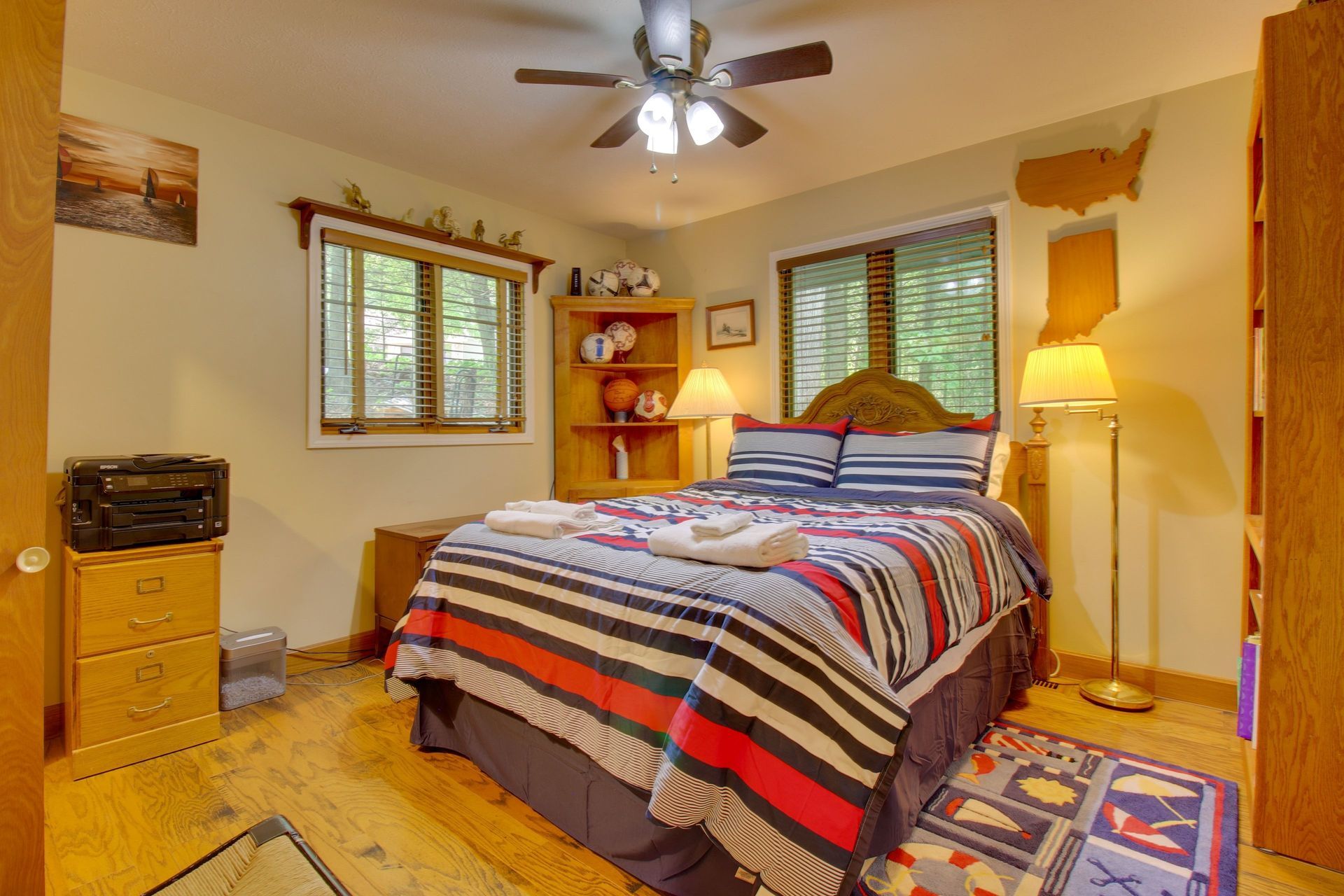 Bedroom with bed, bookshelf, windows, and a ceiling fan. Striped quilt, wooden floor.